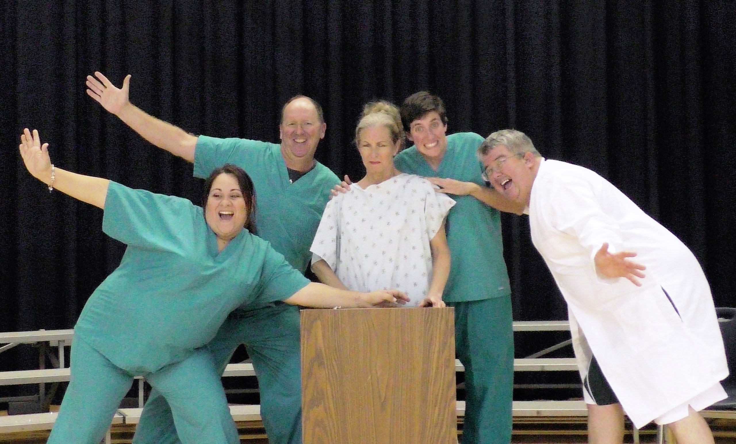 Photo of cast members in scrubs and one in the center in a hospital gown
